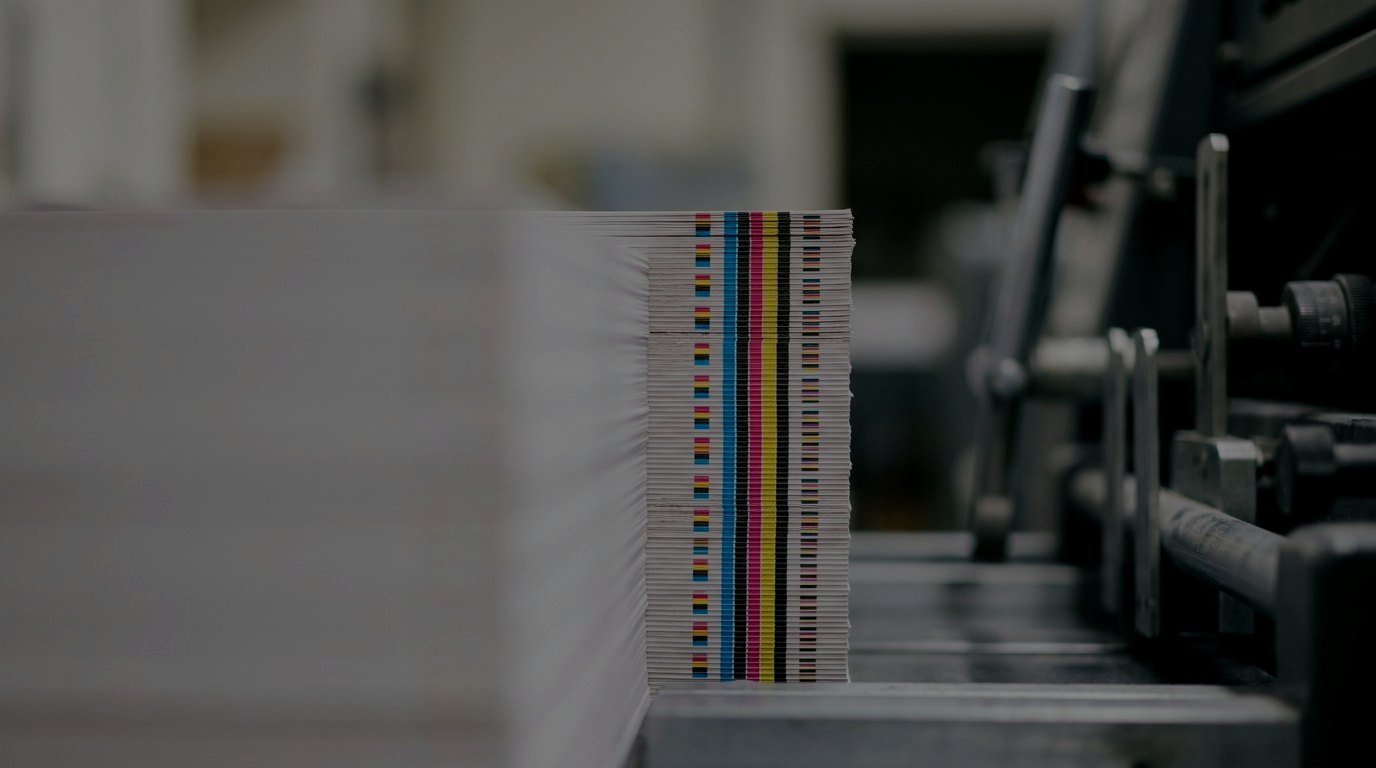 The edge of a fresh stack of printed sheets, its colour bars forming cyan, magenta, yellow and black stripes beside the press.
